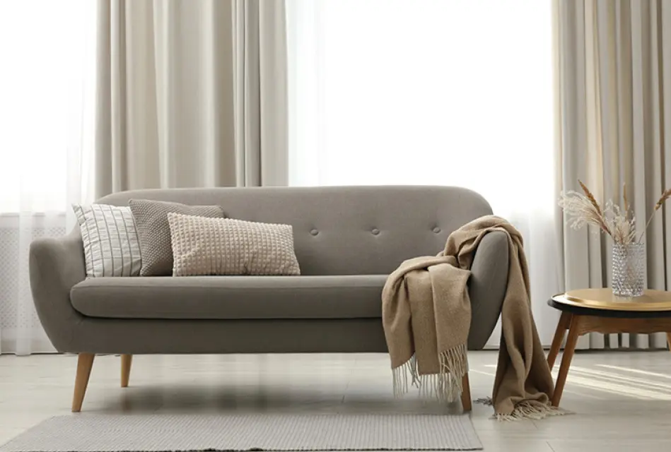 Graues Sofa mit Kissen und Decke vor hohen Vorhängen, runder Beistelltisch mit Vase rechts auf hellem Teppich